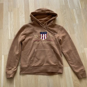 Brun hoodie från GANT - Säljer en snygg brun hoodie från GANT med ett stort tryck på framsidan. Hoodien har en känguruficka och justerbar huva. Perfekt för en avslappnad stil. Tillverkad i polyester för en mjuk och bekväm känsla.