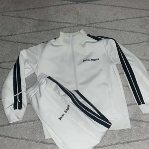 Vit tracksuit från palm angels - Snygg vit träningsjacka från Palm Angels med svarta ränder längs ärmarna och dragkedja framtill. Perfekt för en sportig look eller avslappnad stil. Matchande byxor finns också.