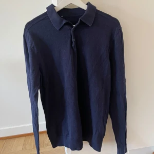 Mörkblå långärmad pikétröja - Säljer en stilren mörkblå pikétröja med långa ärmar och klassisk krage. Perfekt för en avslappnad men ändå uppklädd look. Tröjan har knappar framtill och är tillverkad i ett mjukt material.