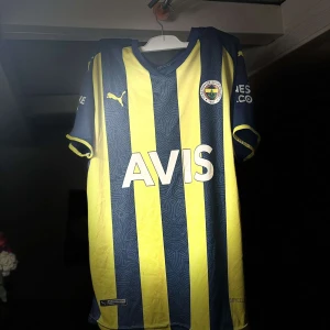 Fenerbache fotbollströja från Puma - Säljer en snygg gul och marinblå randig fotbollströja från Puma. Tröjan har korta ärmar och klubbmärke på bröstet. Perfekt för fotbollsfans som vill visa sitt stöd med stil! 🟡🔵