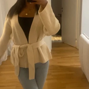 Beige ribbad kofta med bälte, jätte skön och fin. - Säljer en mysig beige ribbad kofta med bälte i midjan. Koftan har långa ärmar och en öppen design som passar perfekt för lager-på-lager-stil. Perfekt för kyliga dagar!