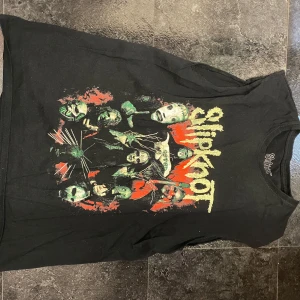 Svart Slipknot t-shirt - Cool svart t-shirt med Slipknot-tryck fram och bak. Framsidan har ett färgstarkt motiv med bandmedlemmarna och logotypen i gult. Baksidan pryds av en röd och orange stjärna med bandets namn. Perfekt för fans av bandet!
