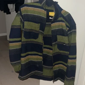 Randig overshirt från carlings - Säljer en cool randig overshirt i grönt och blått med dragkedja framtill. Jackan har en avslappnad passform och är perfekt för lager-på-lager-stil. Den har långa ärmar och en praktisk bröstficka. Köpt från carlings för 899kr och använt fåtal gånger