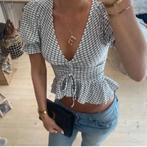 Svartvit rutig blus med volang - Snygg svartvit rutig blus med korta ärmar och volangdetalj i nederkant. Blusen har en v-ringad design och knytning framtill som ger en feminin touch. Perfekt för en stilren look.