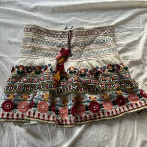 Broderad kjol från Zara - Säljer en vacker broderad kjol från Zara med färgglada blommönster och volanger. Kjolen har en knytning i midjan med tofsar i rött och gult. Perfekt för att ge en bohemisk touch till din outfit!