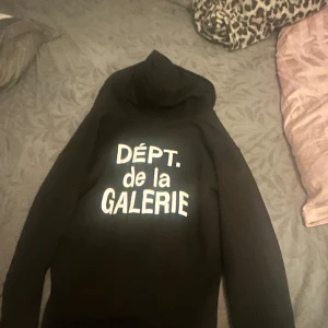 Svart hoodie från gallery dept  - Säljer en svart hoodie med texten 'DÉPT. de la GALERIE' på framsidan. Hoodien har dragkedja och är perfekt för en avslappnad stil. Den är designad av Josue Thomas och har en klassisk passform.