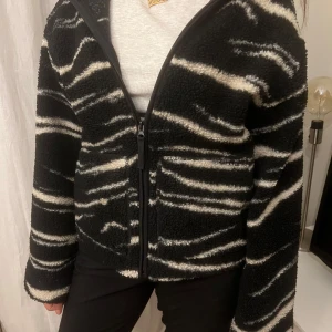 Svart och vit teddyjacka med dragkedja - svart teddyjacka med vita zebramönster och dragkedja framtill. Perfekt för kyliga dagar med sin fluffiga och varma design.