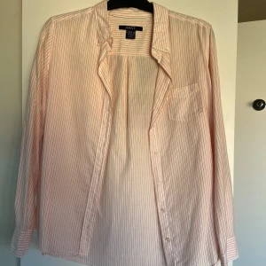Randig skjorta från GANT - Snygg randig skjorta från GANT i vitt och rosa. Skjortan har långa ärmar och en klassisk krage. Den är försedd med knappar framtill och en bröstficka med broderad logotyp. Perfekt för en stilren look. OBS skjortan är inte mörk över ryggen, den är bara upphängd över en tavla vilket syns igenom lite😧