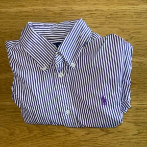 Lila och vit randig skjorta från Ralph Lauren - Säljer en klassisk skjorta från Ralph Lauren med smala lila och vita ränder. Skjortan har lång ärm, knappar framtill och broderad logga på bröstet. Perfekt för dig som gillar stilrena plagg med en twist.
