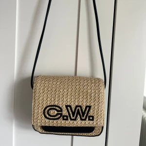 Beige axelväska från Carin Wester - Säljer en beige axelväska med svart C.W.-logga framtill från Carin Wester. Väskan har en flätad struktur och svarta detaljer längs kanterna samt axelrem. Perfekt för dig som vill ha en snygg accessoar med unik design. Skriv för fler bilder💞