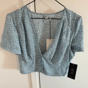 topp  - Söt ljusblå croppad omlottopp från NA-KD med små vita blommor. Toppen har korta ärmar och djup v-ringning. Perfekt för varma dagar och enkel att styla med jeans eller kjol.