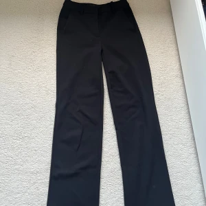 Svarta kostymbyxor från Vero Moda - Klassiska svarta kostymbyxor från Vero Moda med raka, vida ben och hög midja. Byxorna har dragkedja, knapp och hyska i midjan samt bälteshällor. Perfekta för en stilren och tidlös look.