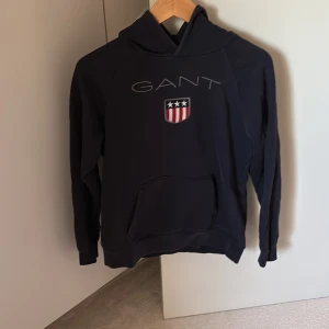 Mörkblå hoodie från GANT - Säljer en mörkblå hoodie från GANT med klassisk logga och sköld på bröstet. Tröjan har huva och en stor magficka framtill. Perfekt för dig som gillar stilrena plagg.