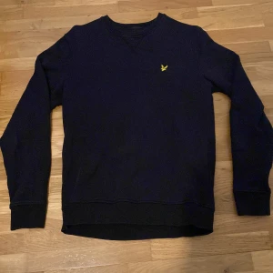 Svart tröja från Lyle & Scott - Säljer en svart tröja från Lyle & Scott med gul broderad logga på bröstet. Tröjan har en rund halsring och långa ärmar. Perfekt för dig som gillar stilrena och klassiska plagg.