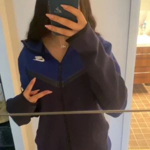 Nike tech  - Mörkblå hoodie från Nike med dragkedja och huva. Jackan har en vit Nike-logga på bröstet och svarta detaljer längs bröstet. Perfekt för en sportig och avslappnad stil.