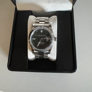 Seiko Wimbledon - Säljer en klassisk Seiko Wimbledon Datejust med svart urtavla och romerska siffror. Klockan har en räfflad bezel och länkarmband i silverfärgat stål. Datumvisning vid klockan tre och automatisk urverk synligt på baksidan.
