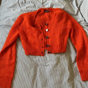 Orange croppad kofta från Divided - Säljer en stickad orange kofta från Divided. Modellen är croppad med långa ärmar och har hjärtformade knappar framtill. Perfekt för att liva upp din outfit med en färgklick! Passar Xs-mindre M