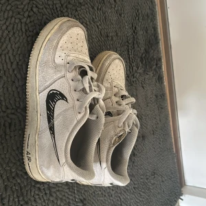 Nike Air Force 1 vita sneakers med svart swoosh - Säljer ett par klassiska Nike Air Force 1 sneakers i vitt med svart handritad swoosh. Skorna har snörning, rund tå och platt sula. Perfekta för dig som gillar streetstyle och vill ha en unik touch på dina sneakers.