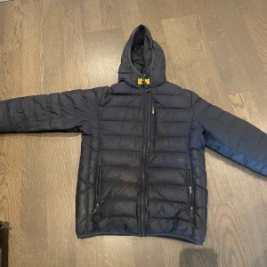 Mörkblå dunjacka från Parajumpers - Svart dunjacka från Parajumpers med huva och dragkedja framtill. Jackan har två sidofickor med dragkedja och en bröstficka. Lättviktig modell med quiltad design och klassisk Parajumpers-detalj vid halsen. Det finns tecken på användning som tex lagningslappar inget man ser vanligtvis, och sen är de någon typ av sol blekning på magen kanske kan gå att få bort är inte säker. Annars är de en fin jacka i ordentligt skick nypris ca 6000kr
