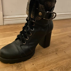 Svarta boots med klack och snörning - Säljer ett par svarta boots från Guess med hög klack, snörning framtill och dekorativt spänne upptill. 