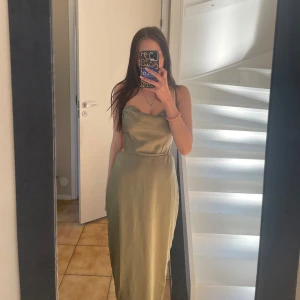 Olivgrön långklänning med öppen rygg - Säljer en olivgrön långklänning med draperad urringning och smala axelband. Klänningen har en elegant öppen rygg och är tillverkad i ett glansigt material som ger en lyxig känsla. Perfekt för speciella tillfällen.