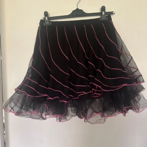 Svart tyllkjol med rosa kant - En fluffig svart tyllkjol med flera lager och vågiga rosa kanter. Kjolen har en voluminös form och är perfekt för dig som vill sticka ut. Resår i midjan för bekväm passform. Helt ny. Onesize. Resår i midjan,