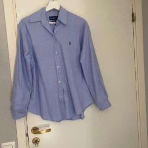 Ljusblå skjorta från Ralph Lauren - Klassisk ljusblå skjorta från Ralph Lauren med broderad logga på bröstet. Skjortan har lång ärm, knappar framtill och är tillverkad i 100% bomull. Perfekt för en stilren och tidlös look.