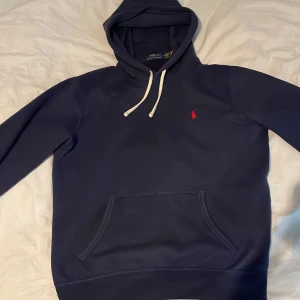 Mörkblå hoodie från Polo Ralph Lauren - Säljer en mörkblå hoodie från Polo Ralph Lauren med vit dragsko och klassisk röd logga på bröstet. Tröjan har magficka och mjukt material, perfekt för en avslappnad stil.