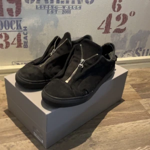 Svarta sneakers med dragkedja - Svarta sneakers i mocka med cool dragkedjedetalj framtill istället för klassisk snörning. Skorna har en rund tå och platt sula, vilket ger en avslappnad och modern look.