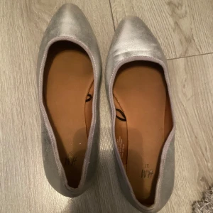 Silvriga ballerinaskor från H&M - Säljer ett par silvriga ballerinaskor från H&M i storlek 37. Skorna har rund tå och är tillverkade i ett glansigt syntetmaterial som ger en snygg metallic-look. Perfekta att styla till olika outfits!