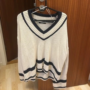 Vit stickad v-ringad tröja med marinblå ränder - Säljer en vit stickad tröja från Gina Tricot med bred v-ringning och marinblå ränder vid hals, ärmslut och nederkant. Tröjan har en avslappnad passform och långa ärmar. Perfekt för lager-på-lager-stil. Köpt för 399kr