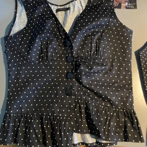 Svart prickig blus med volang - Svart blus med vita prickar från Gina Tricot. Blusen har stora knappar framtill, v-ringning och en volang nedtill. Ärmlös modell som passar perfekt till sommaren.