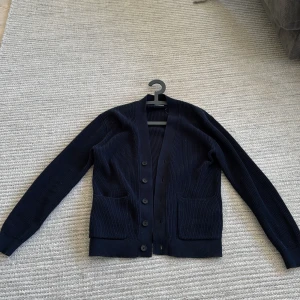 Mörkblå stickad kofta med knappar - Säljer en klassisk mörkblå stickad kofta med v-ringning och stora framfickor. Koftan har långa ärmar och stängs med fem knappar framtill. Perfekt för lager-på-lager och enkel att matcha med olika outfits. Bara använd 1 gång passar 15/16 åringar 
