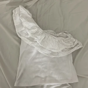 Vit oneshoulder topp med volang från Gina Tricot - Superfin vit oneshoulder topp från Gina Tricot med bred volang i broderad spets. Toppen är ribbad och har en snygg asymmetrisk design som gör den extra speciell.