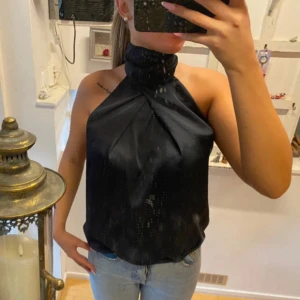 Mörkblå  halterneck-top från Zara Basic - Säljer en mörkblå halterneck-top från Zara Basic med hög krage och elegant omlott-drapering framtill. Toppen är ärmlös och har en snygg, glansig finish som ger en lyxig känsla. Perfekt att styla med jeans eller kjol.