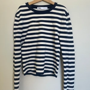 Randig långärmad tröja från Zara - Säljer en klassisk långärmad tröja från Zara med marinblå och vita horisontella ränder. Tröjan har rund halsringning och är tillverkad i mjukt material, perfekt för en stilren vardagslook. När jag strök tröjan råkade jag smälta den lite i fram, därav priset. Detta är inget som syns på långt håll. Kan skicka närmare bild på skadan☺️
