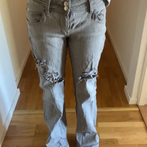 Grå jeans med slitningar - Säljer ett par grå jeans med slitna detaljer på båda benen. Byxorna har normal passform, stängs med dragkedja och knappar. Perfekta för en avslappnad och trendig look.