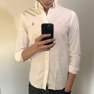 Vit skjorta från Ralph Lauren - Klassisk vit skjorta från Ralph Lauren med broderad logga på bröstet. Skjortan har lång ärm, knappar framtill och krage med knappar. Perfekt för en clean och stilren look. Storlek S slimfit 