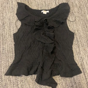 Svart volangtopp  - Säljer en jättefin svart ärmlös topp från Gina Tricot med volangdetaljer längs halsringningen och nederkanten. Den är aldrig använd💗😊