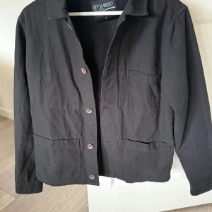 Svart jacka från Key Largo - Säljer en svart overshirt från Key Largo i 100% bomull. Jackan har knappar framtill, klassisk krage och tre fickor på framsidan. Tillkommer en extra knapp. 