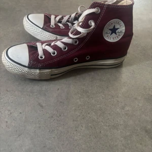 Vinröda Converse All Star high-top sneakers - Klassiska vinröda Converse All Star high-top sneakers med vit sula och vita snören. Skorna har rund tå, platt sula och tydlig Converse-logga på sidan. Perfekta för en avslappnad och stilren look.