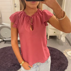 Rosa blus med volangärm och knyt - Söt rosa blus med korta volangärmar och knytning vid halsen. Blusen har en liten öppning framtill och är tillverkad i ett mjukt, lätt tyg. Perfekt för vår och sommar.