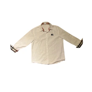 Vit skjorta från Burberry med rutiga detaljer - Säljer en klassisk vit skjorta från Burberry med ikoniska rutiga detaljer på insidan av kragen och ärmsluten. Skjortan har lång ärm, knappar framtill och broderad logga på bröstet. Perfekt för dig som gillar stilrena plagg med lyxig känsla.