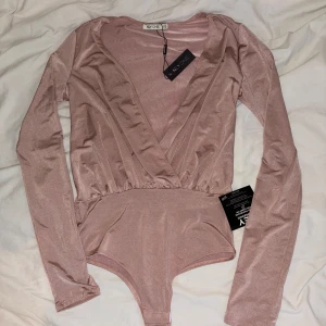 Body från Nelly - Säljer en ljusrosa bodysuit från NLY One med omlott-design och djup v-ringning, oanvänd.