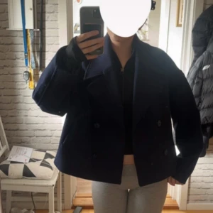 Mörkblå croppad kavajjacka från Lager 157 - Säljer en mörkblå croppad kavajjacka med dubbelknäppning och bred krage. Jackan har långa ärmar och är tillverkad i ett tjockare material som känns lite ulligt. Perfekt för dig som gillar en stilren och trendig look.