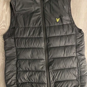 Svart dunväst från Lyle & Scott - Svart dunväst med dragkedja framtill och gul broderad logotyp på bröstet. Västen har quiltad design och står för en klassisk stil. Perfekt att bära över en hoodie eller tröja.