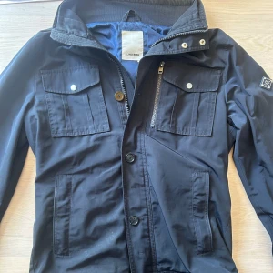 J Lindberg jacka - Säljer en svart jacka från j Lindberg med dragkedja och knappar framtill. Jackan har hög krage, långa ärmar och diskret logga på ärmen. Perfekt för blåsiga dagar och enkel att matcha med det mesta.