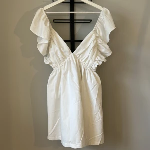 Vit klänning  - Superfin vit kortklänning med djup v-ringning och volangdetaljer längs axelbanden. Klänningen har knytning i ryggen och är ärmlös, perfekt till studenten. Endast testad aldrig använd, prislapp kvar. 