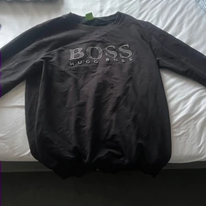 Svart sweatshirt från Hugo Boss - Säljer en svart sweatshirt från Hugo Boss med stort broderat BOSS-tryck på bröstet. Tröjan har rund halsringning och långa ärmar. Perfekt för en stilren och avslappnad look.