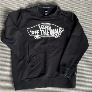 Vans - Svart sweatshirt - Säljer en svart långärmad sweatshirt från Vans med stort vitt 'Off The Wall'-tryck på bröstet. Tröjan har rund halsringning och ribbade muddar vid ärmslut och nederkant. Perfekt för en avslappnad streetstil. Storlek M (ungdom). 